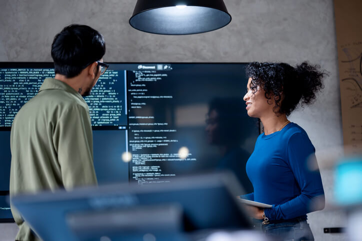 Two software developers discuss a coding project displayed on a TV screen in a tech business office.